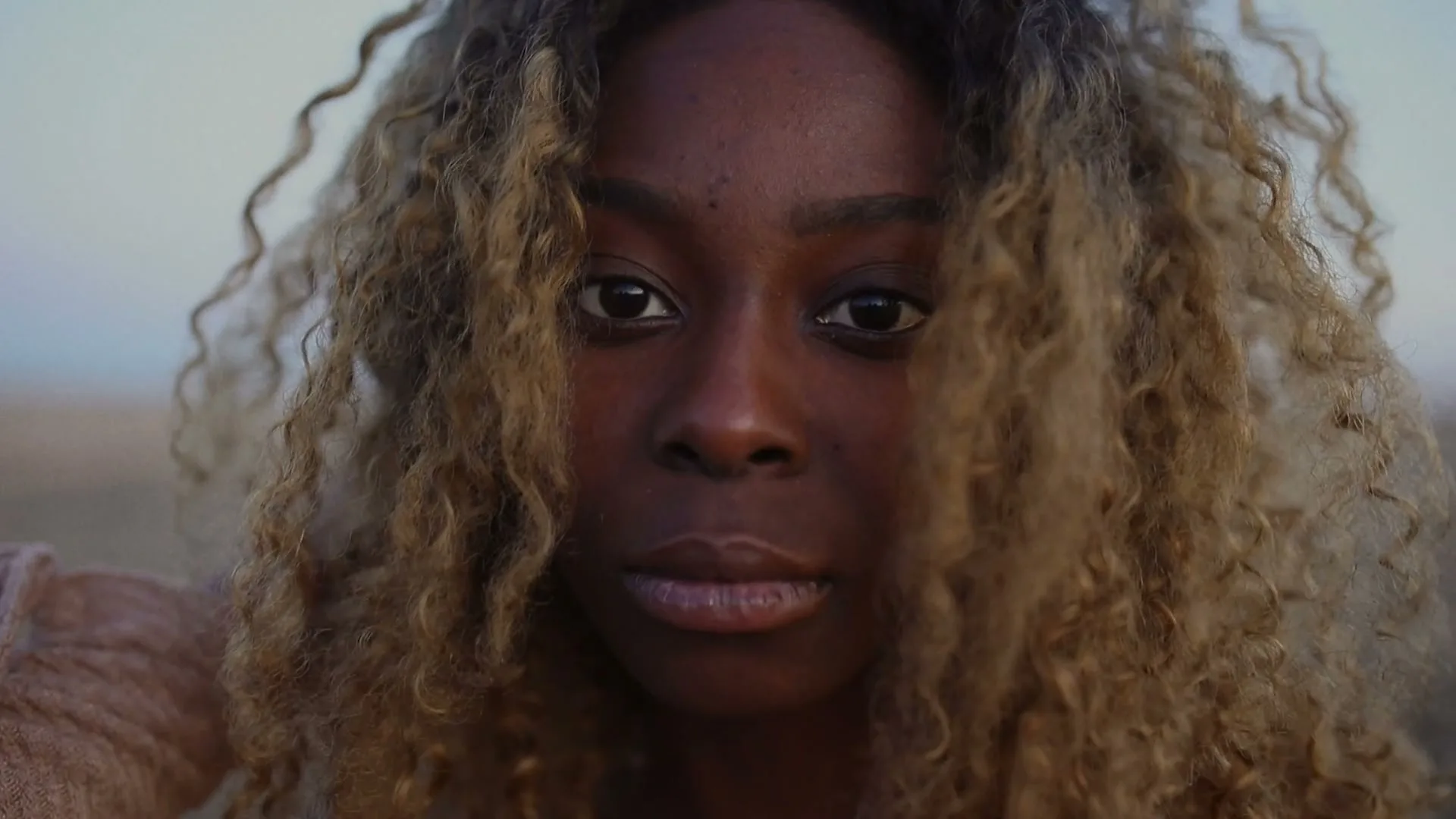 A young Black woman with curly blonde hair looks directly at the camera in an outdoor setting during golden hour.