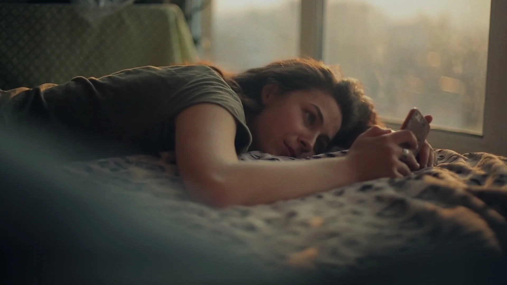 A young woman with curly hair lies on a bed, looking at her phone with a soft smile, bathed in golden hour light from a window.