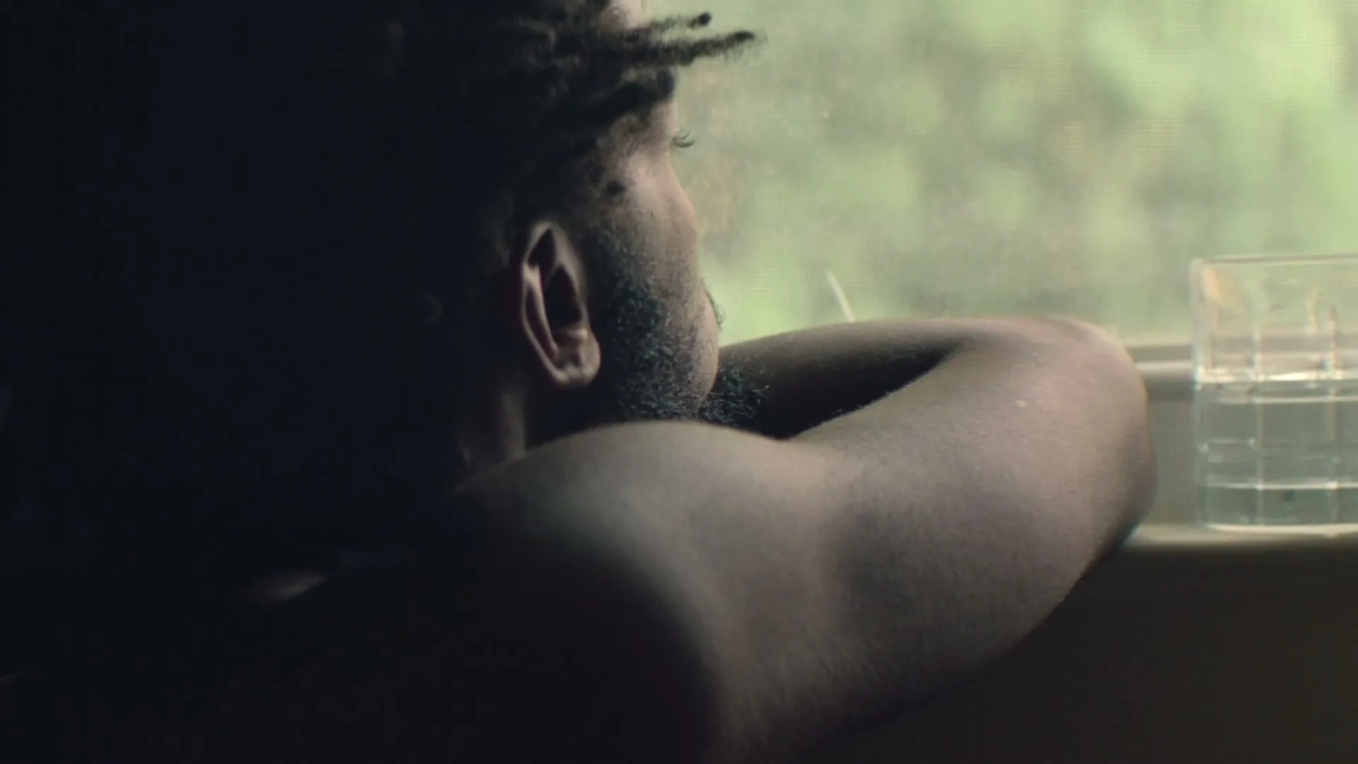A man with dreadlocks and a beard, shirtless, leans on a windowsill looking out at green foliage, with a glass of water nearby.