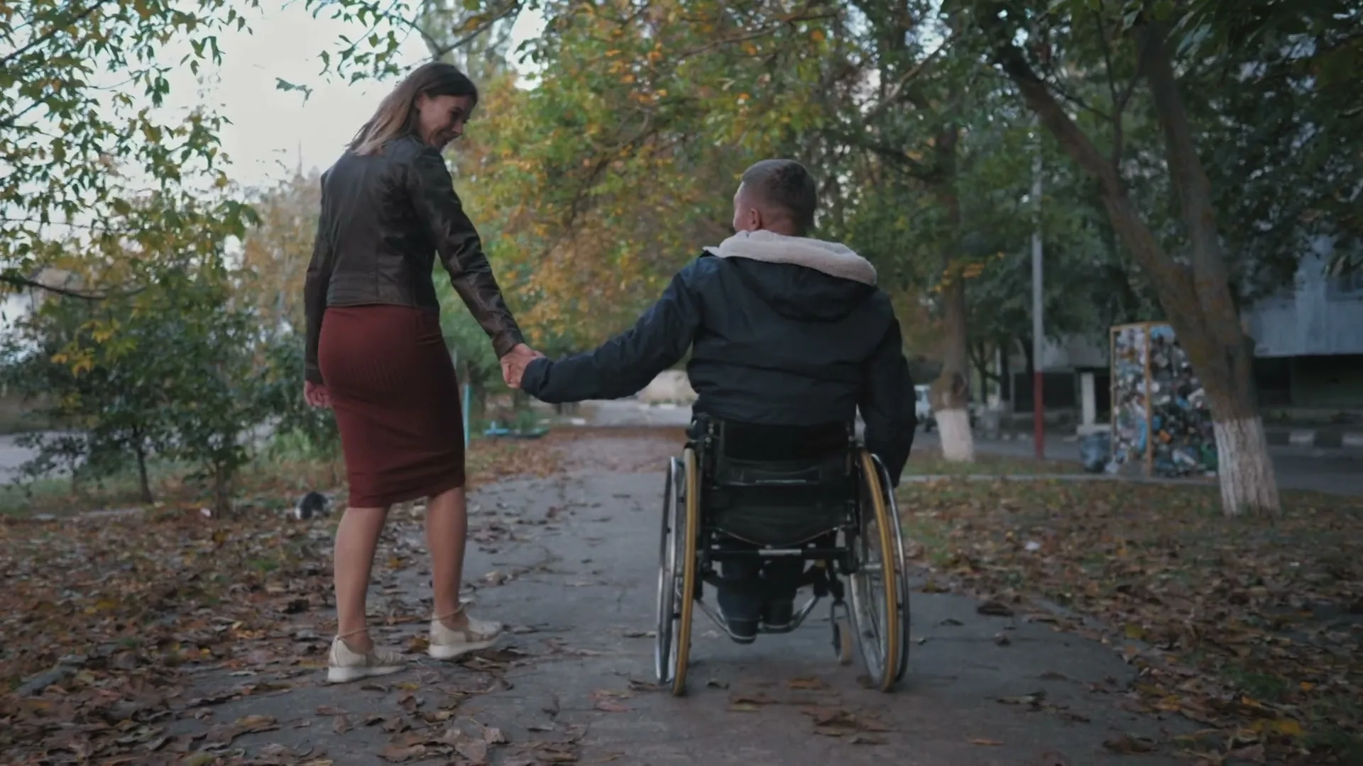 A woman in a dark jacket and red skirt holds hands with a man in a wheelchair, as they move along a path with fallen leaves in an outdoor setting.