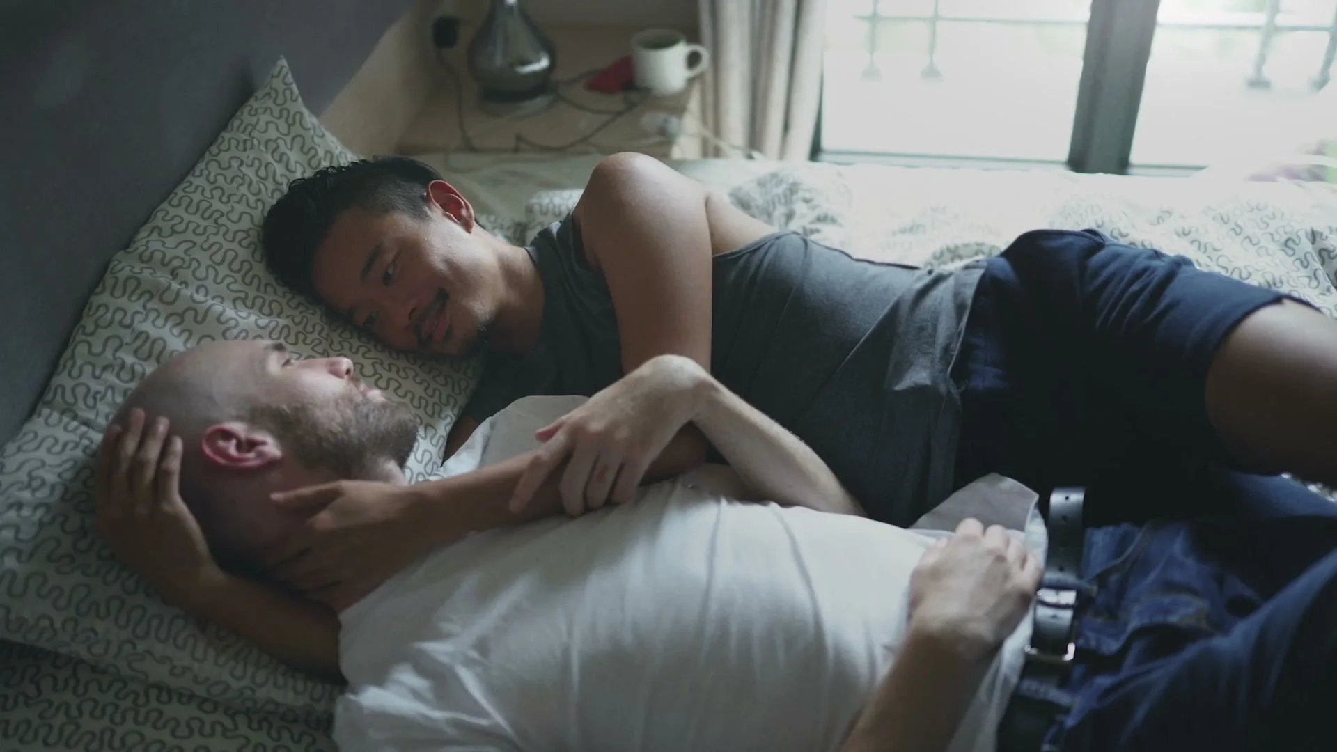 Two men lie in bed, one with a shaved head and white t-shirt, the other with dark hair and a grey tank top, looking at each other intimately.