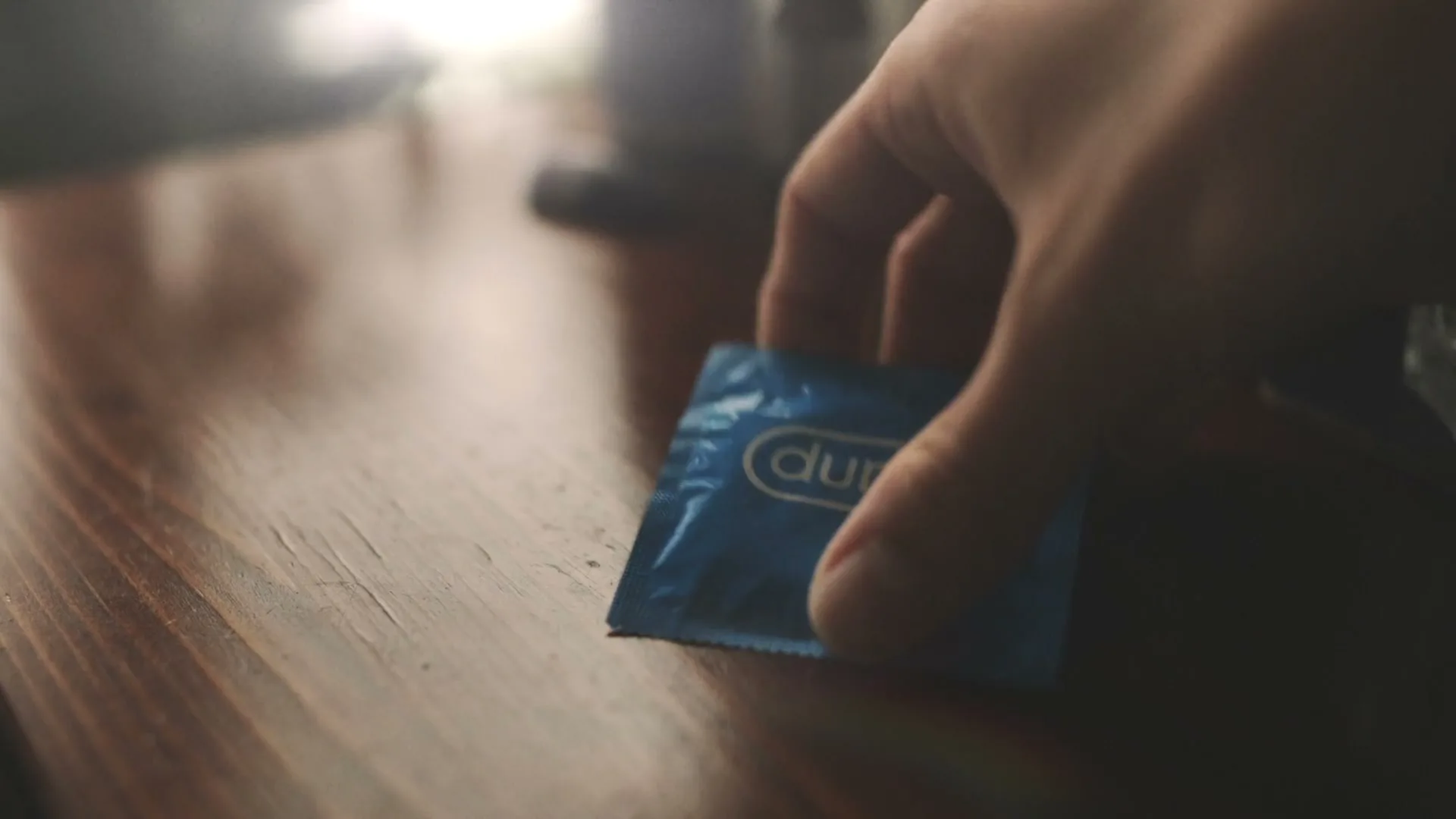 A hand reaches into frame to pick up a Durex condom wrapper from a wooden table.