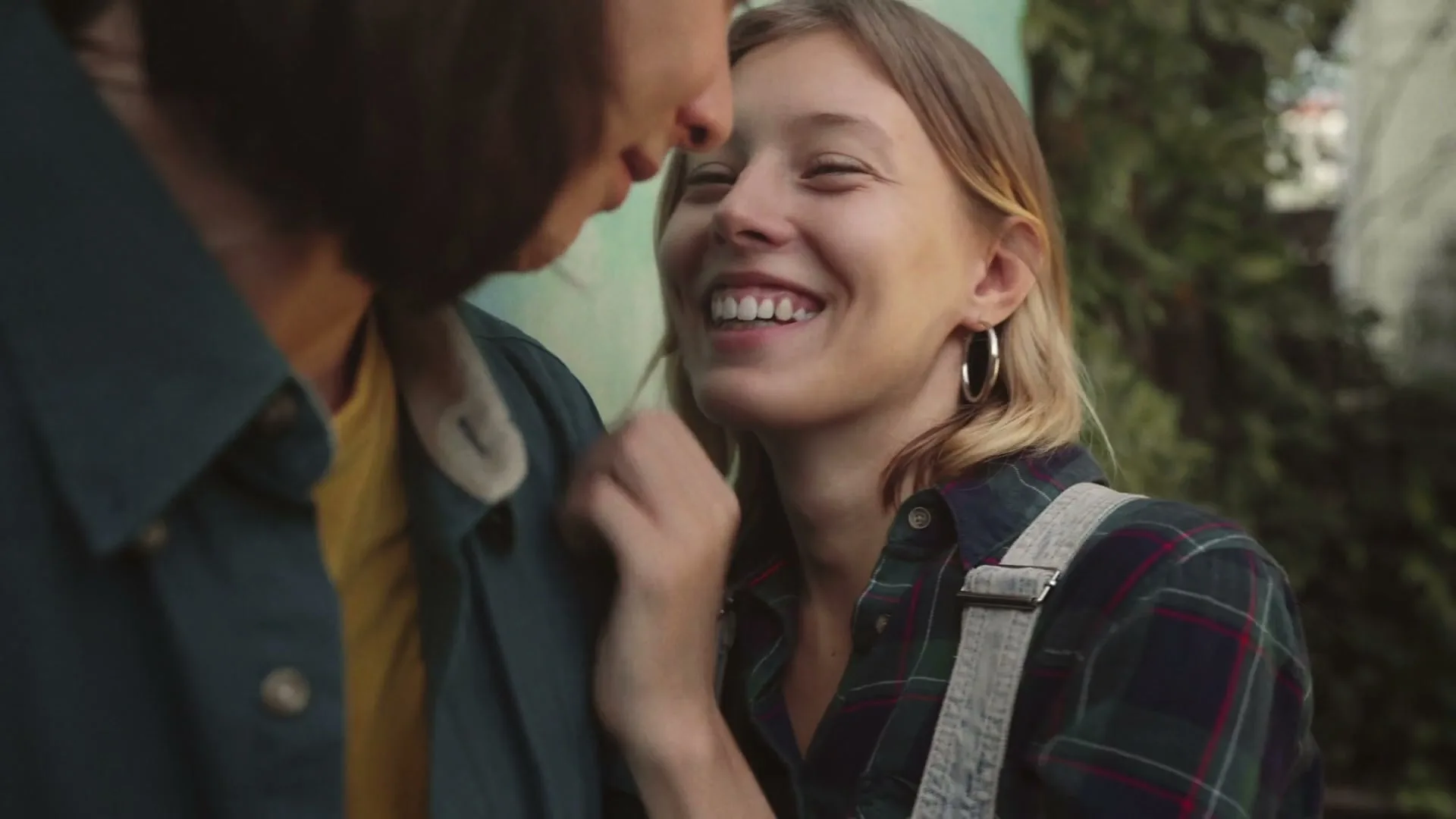 A young woman with blonde hair and hoop earrings smiles at a person with dark hair, touching their face, in an outdoor setting.