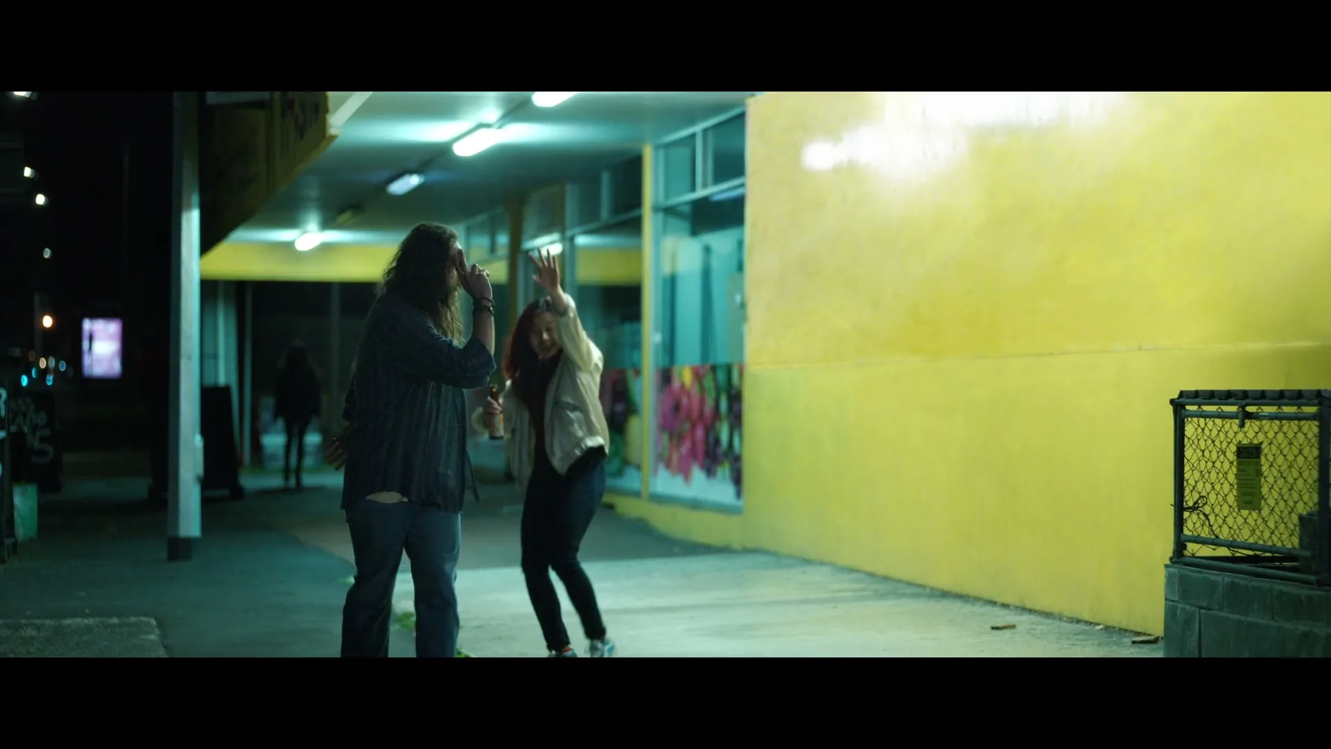 A young man with long hair and a young woman are playfully pushing each other on a brightly lit sidewalk at night.