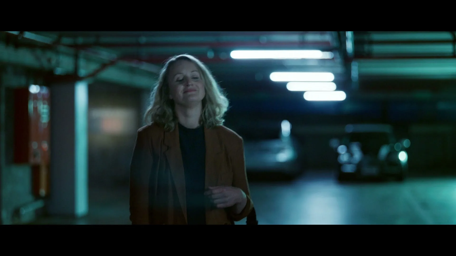 A woman with blonde hair and a brown jacket walks through a dimly lit parking garage, smiling with her eyes closed.