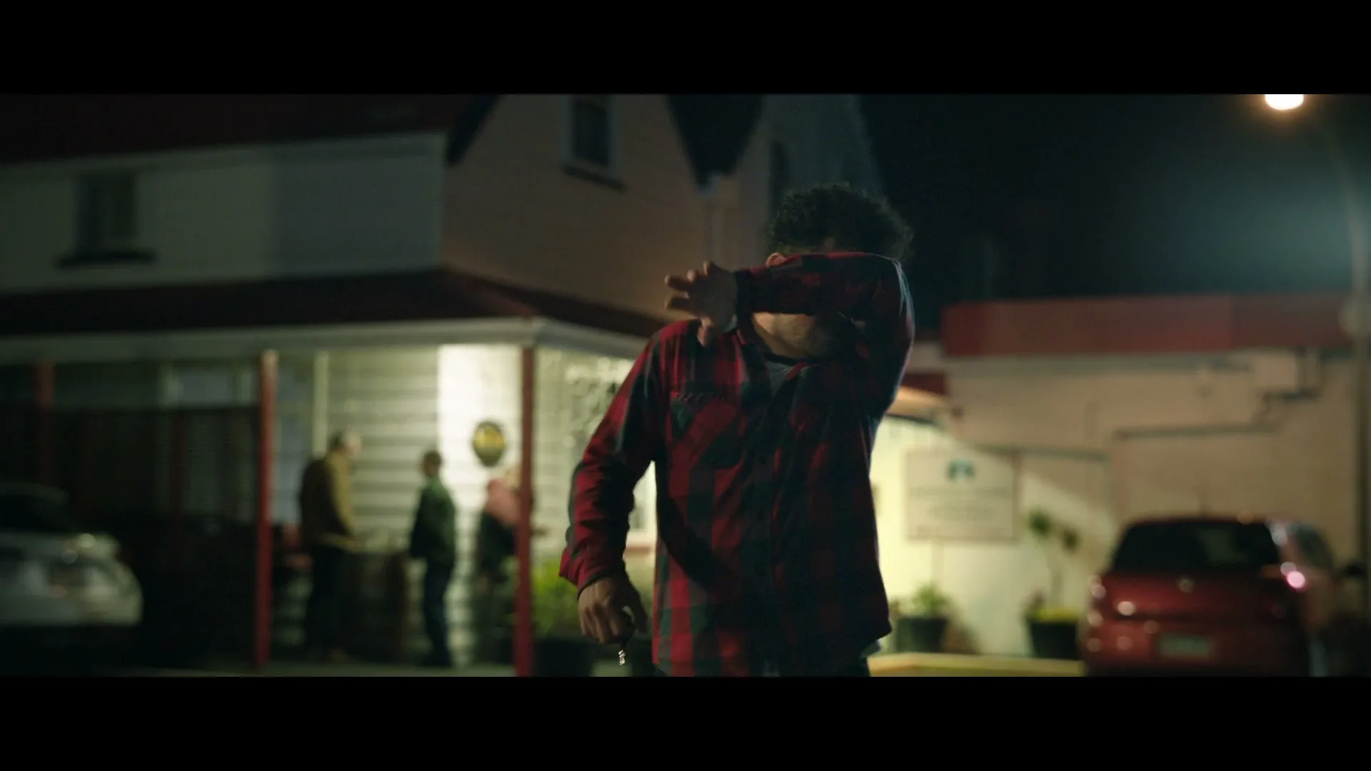 A man in a red plaid shirt stumbles out of a building at night, shielding his eyes from the light, then looks down at his hands.