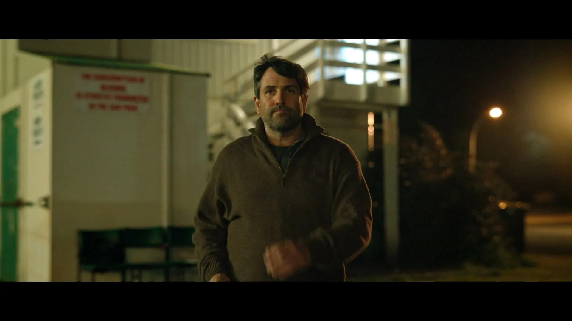 A man with a beard and a brown sweater walks towards the camera at night, with a building and streetlights in the background.