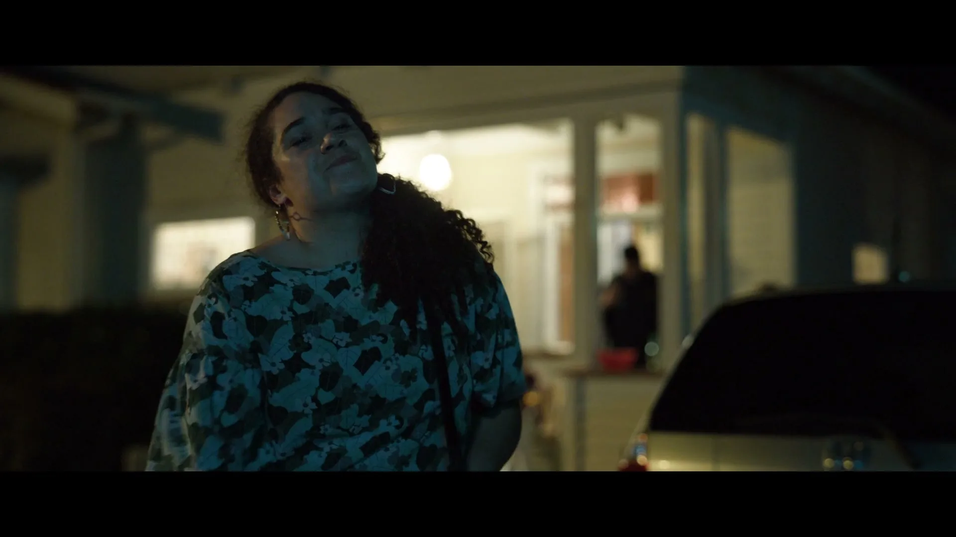 A woman with curly hair and a patterned top stands outside a house at night, looking up with a pained expression.