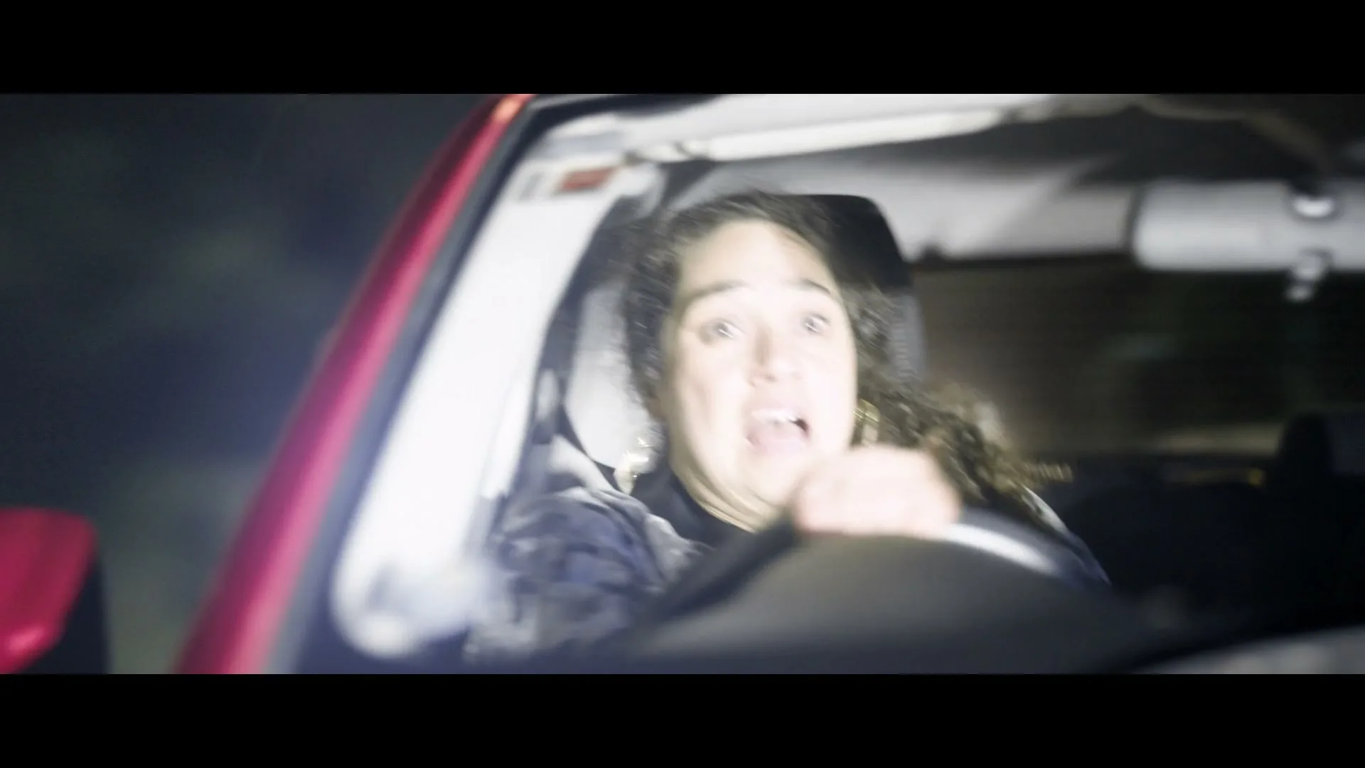 A woman with curly hair and gold earrings drives a red car at night, looking shocked and scared.