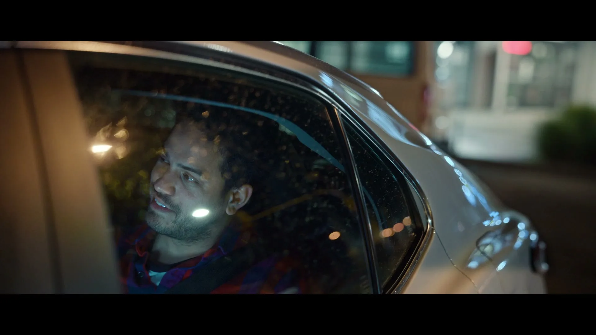 A man with dark hair and a beard, wearing a plaid shirt, looks out the window of a car at night, smiling slightly.