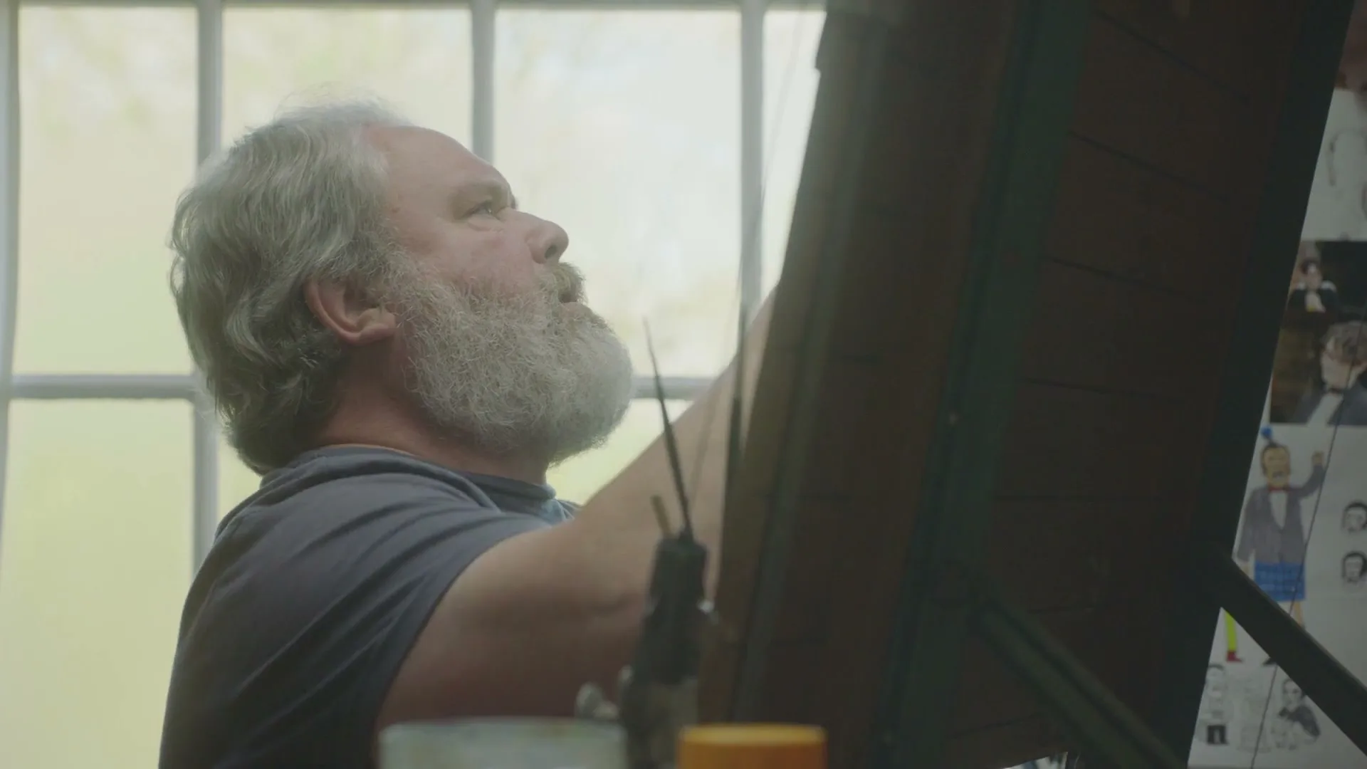 A man with a white beard and grey t-shirt is painting on an easel in front of a window, looking up at his work.
