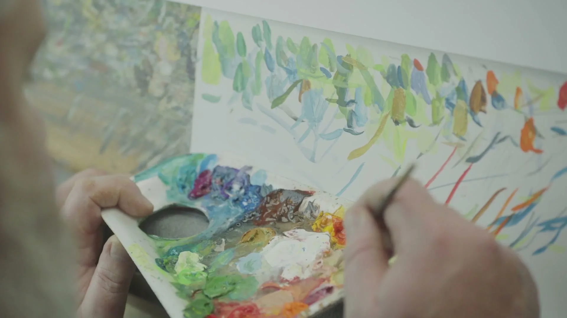 An older person's hands hold a paint palette and brush, adding dabs of color to a vibrant, abstract painting on a canvas.