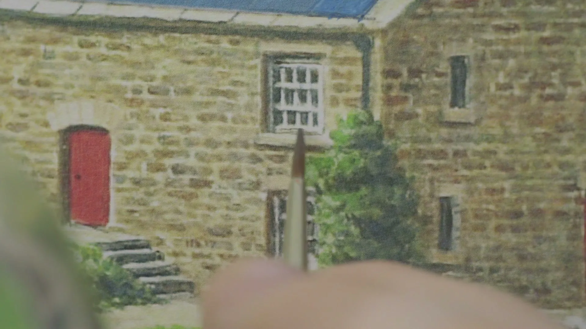 A person's hand holds a paintbrush, adding details to a miniature painting of a stone house with a red door and white windows.