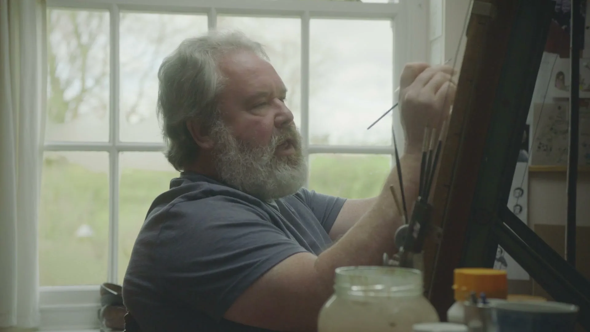 A man with a white beard and gray t-shirt paints at an easel by a window in a bright room.
