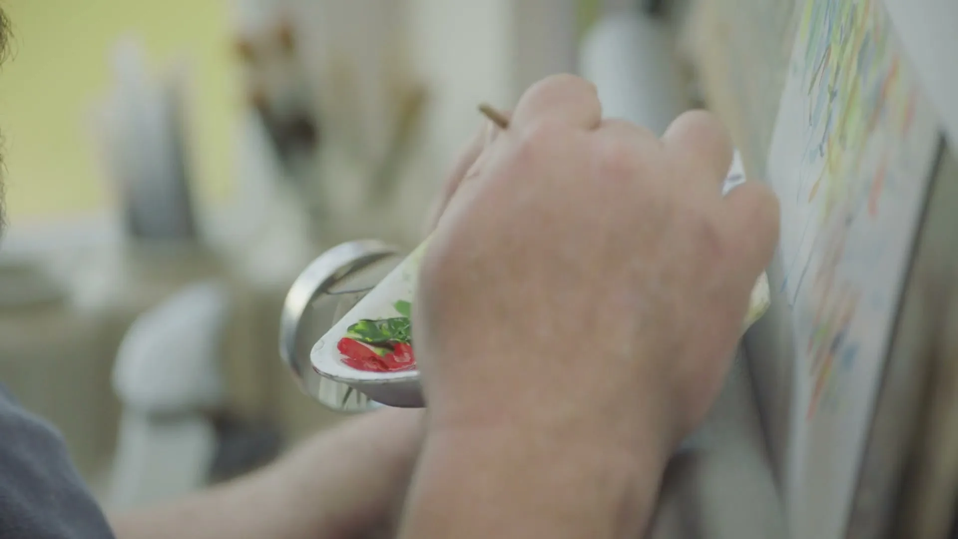 A person's hands hold a paint palette and brush, mixing colors while painting on a canvas in a bright, natural setting.