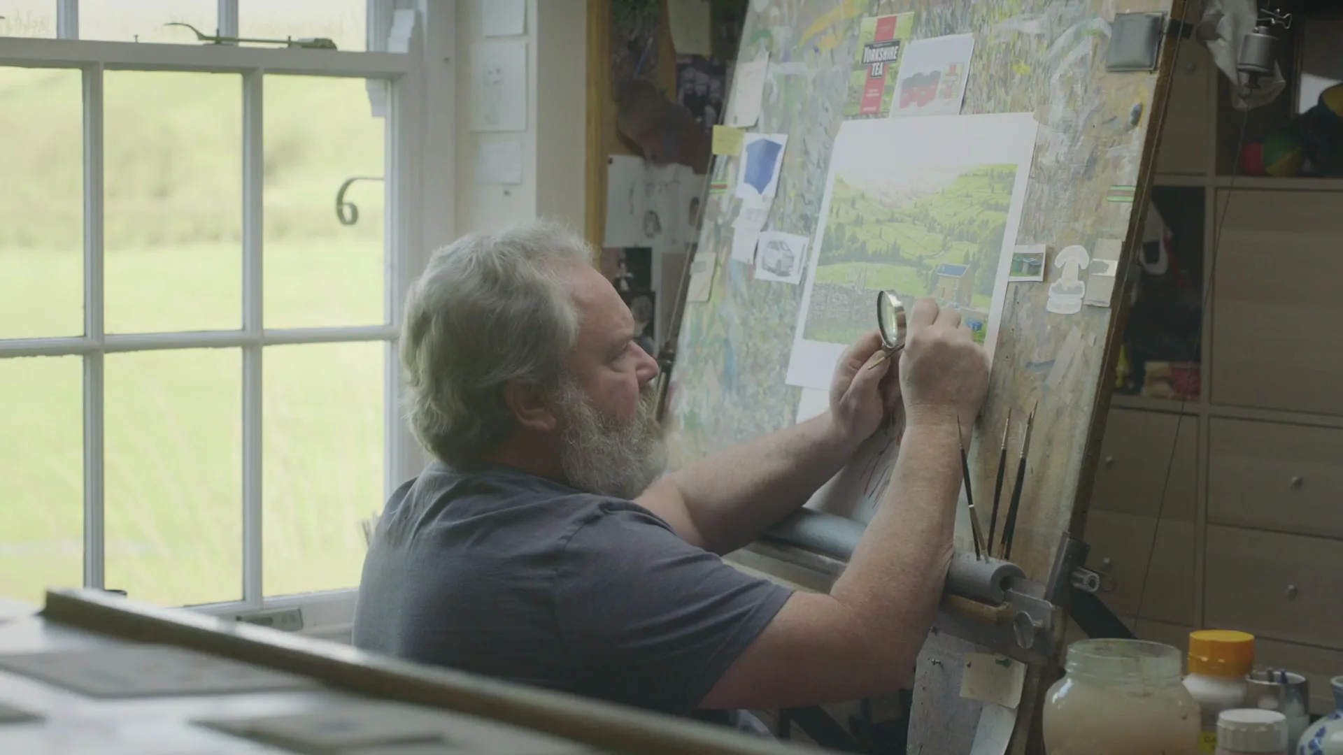 A middle-aged man with a grey beard and t-shirt uses a magnifying glass to examine a detailed drawing of a landscape on an easel in a sunlit studio.