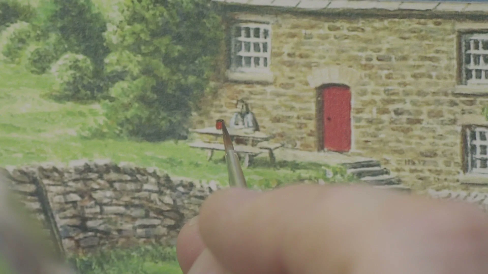 A hand holds a paintbrush, adding detail to a watercolor painting of a stone house with a red door and a person sitting at a picnic table outside.