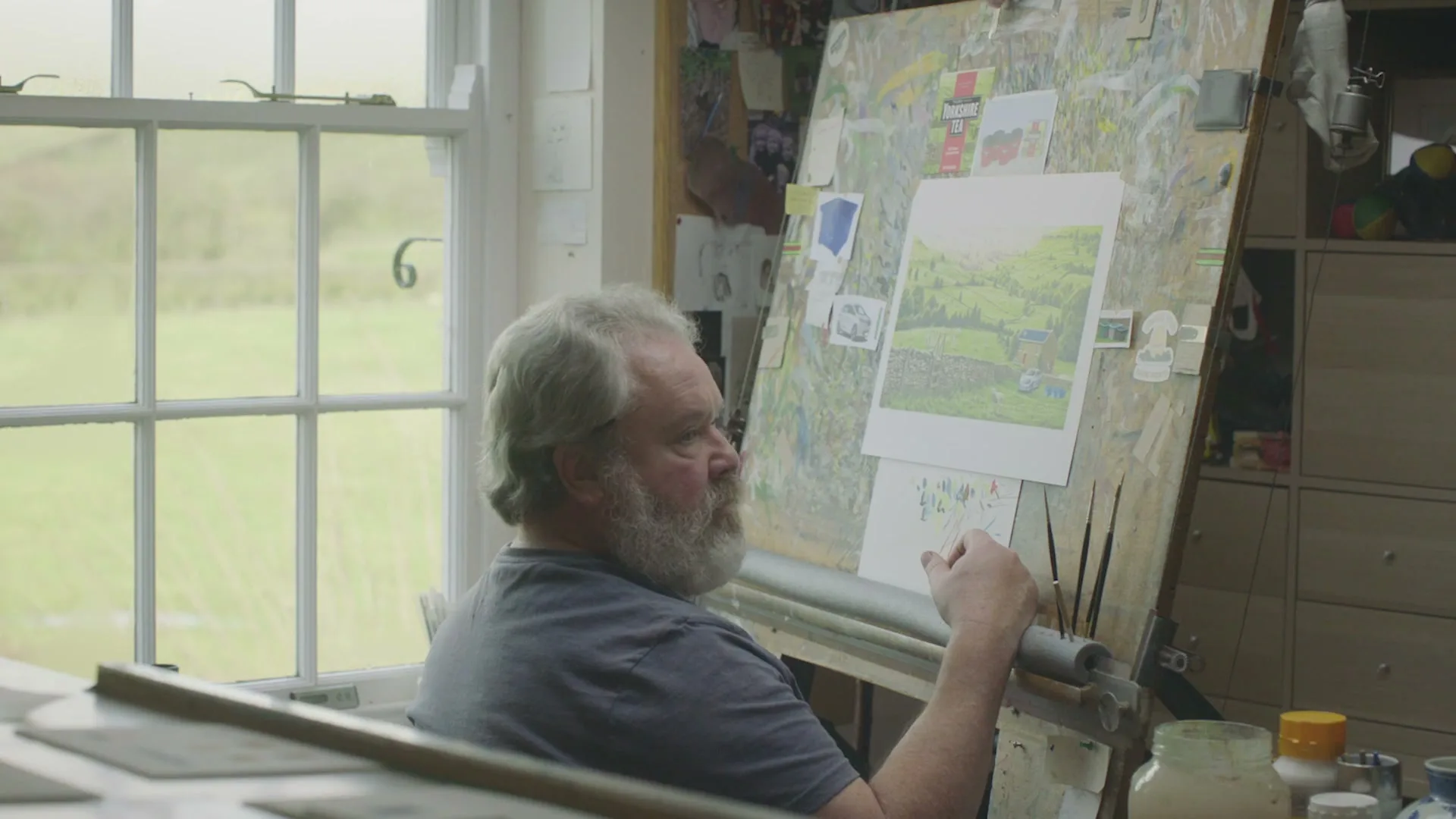 A middle-aged man with a grey beard sits at an easel, painting a landscape, in a sunlit art studio.
