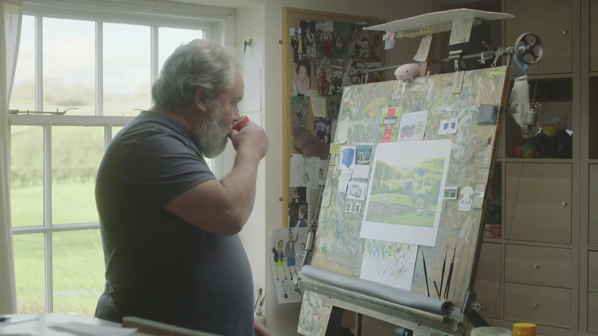A middle-aged man with a beard, wearing a grey t-shirt, sips from a red mug while looking at a painting on an easel in a sunlit room.