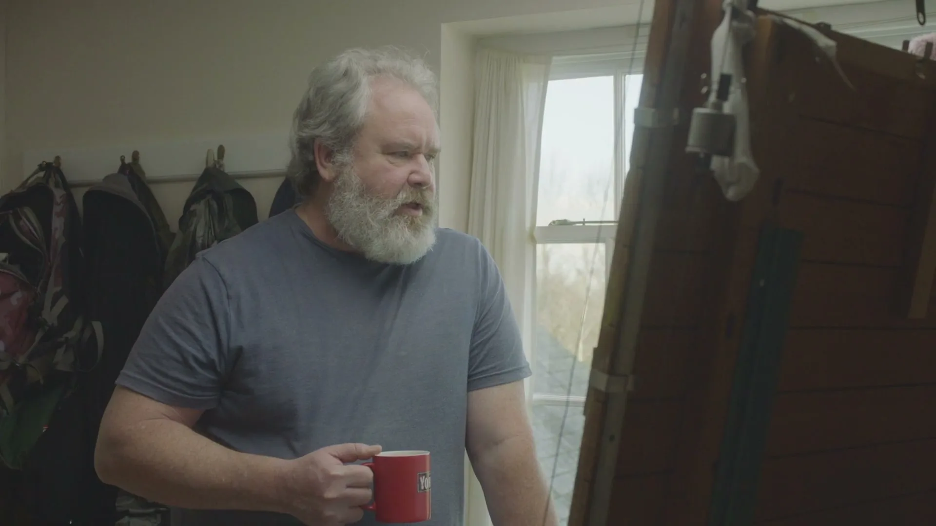 A man with a white beard holds a red mug and looks intently at a wooden structure in a room with a window.