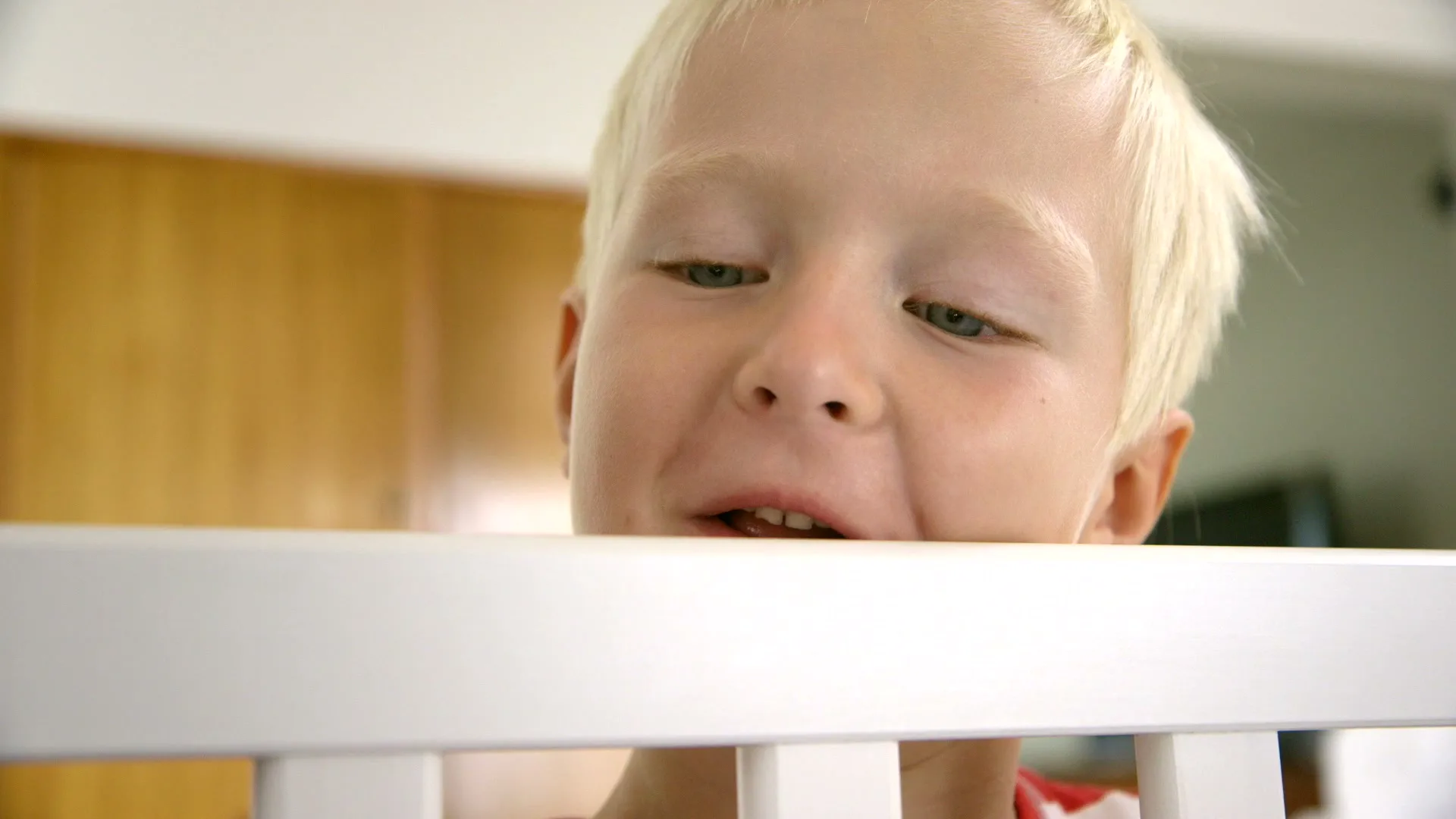 A young blonde boy with blue eyes peeks over the top of a white crib, looking down with a slight smile in a bright room.