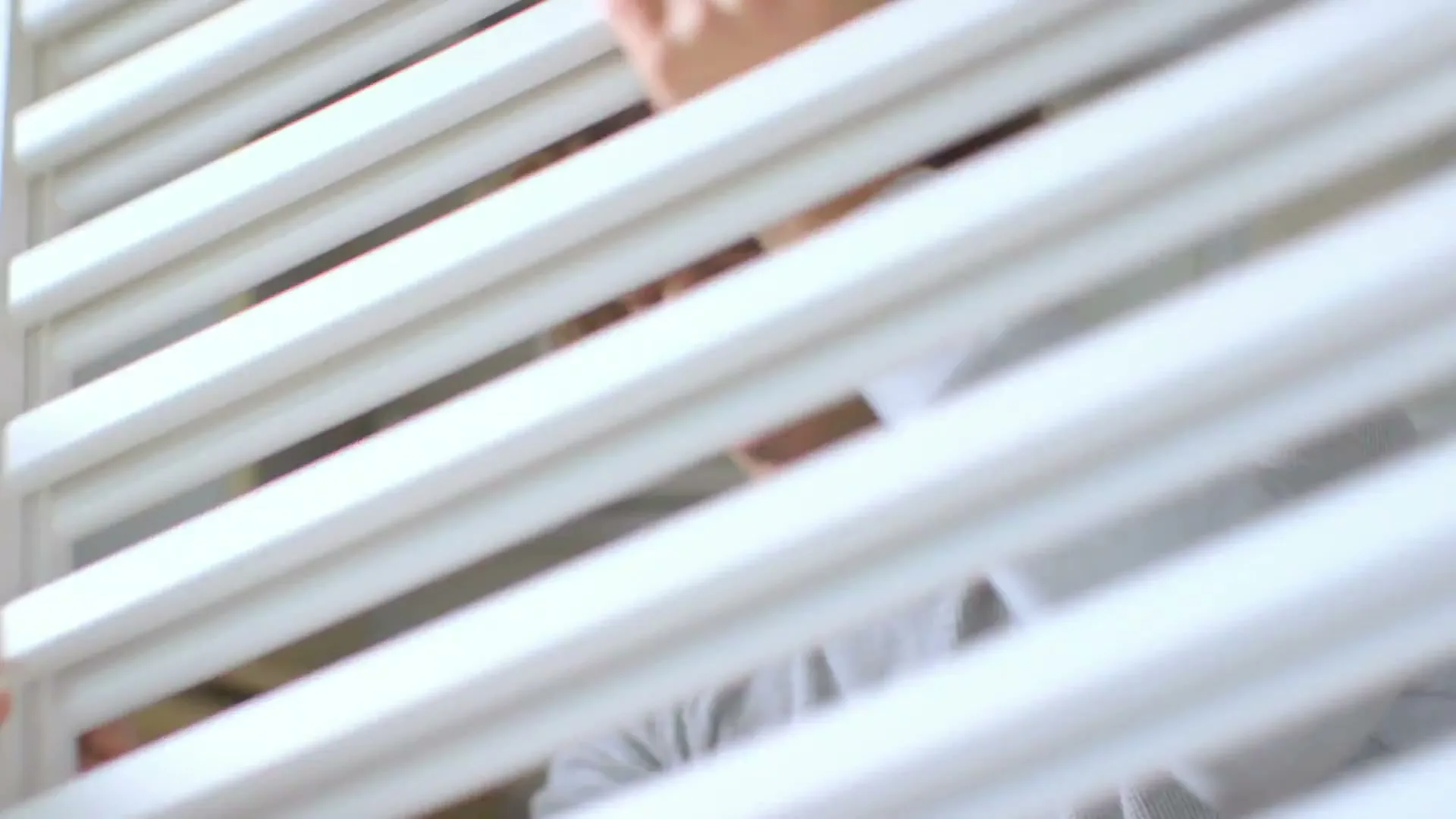 A man with curly brown hair, wearing a grey shirt, assembles a white baby cot in a bright room.