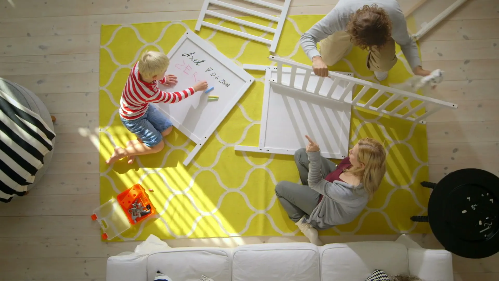A family of three, a man, woman, and child, are assembling a white crib on a yellow rug in a bright living room, with the child drawing on a whiteboard.