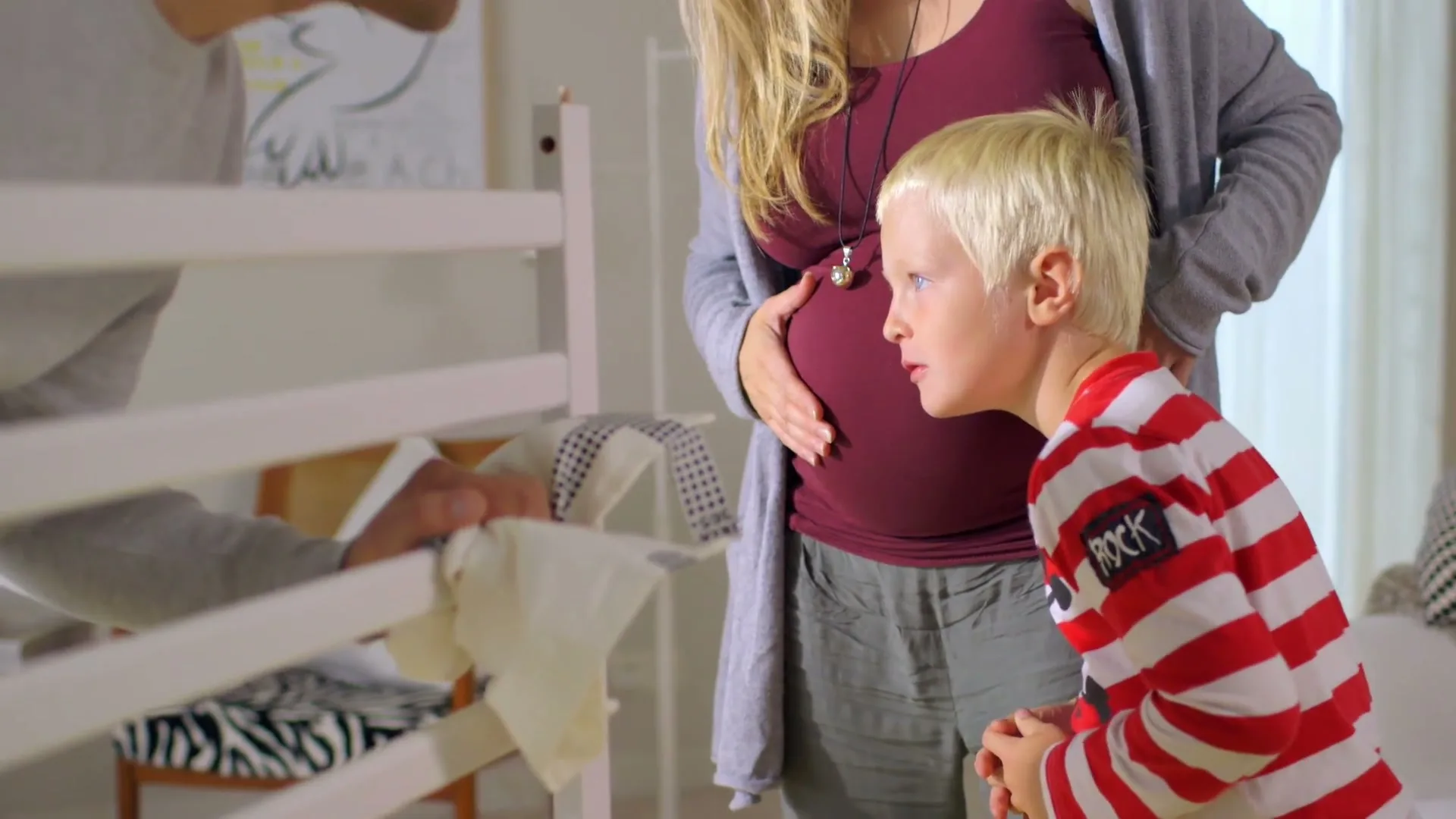 A pregnant woman, a man, and a young boy are in a nursery, looking at a baby cot, with the woman holding her belly.