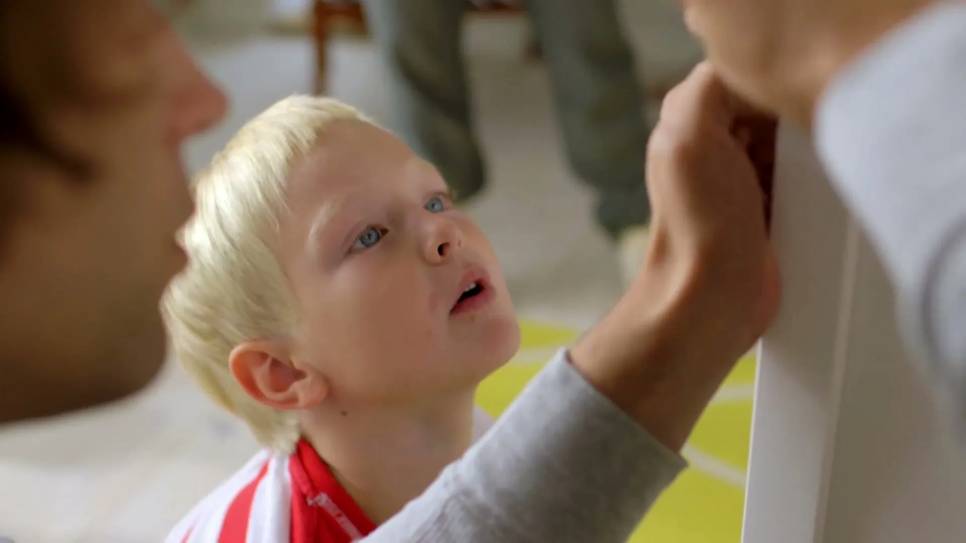 A young boy with blonde hair looks up intently as an adult's hand works on assembling furniture in a bright room.