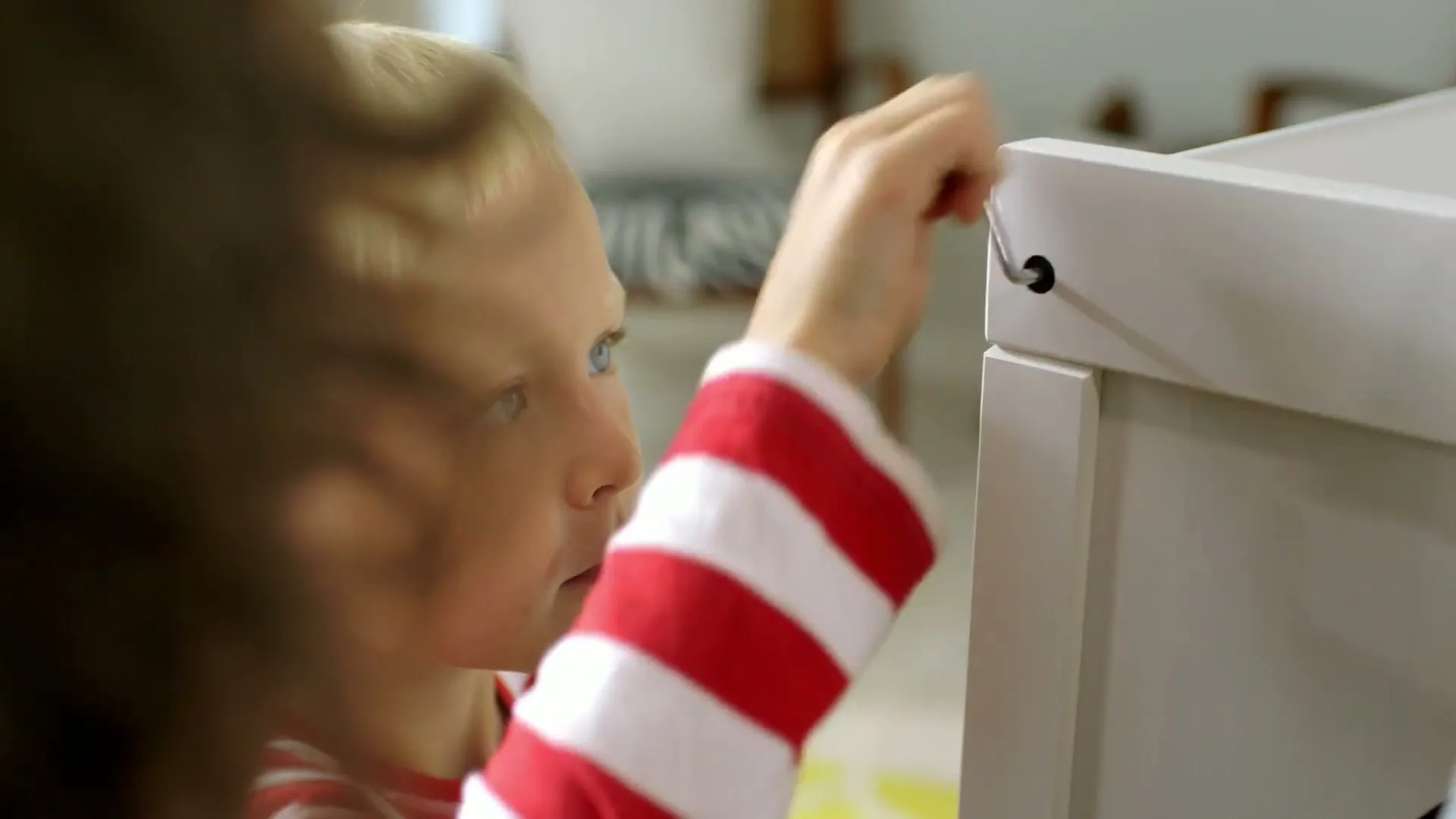 A young boy with blonde hair and a red and white striped shirt helps an adult assemble a white piece of furniture in a bright room.
