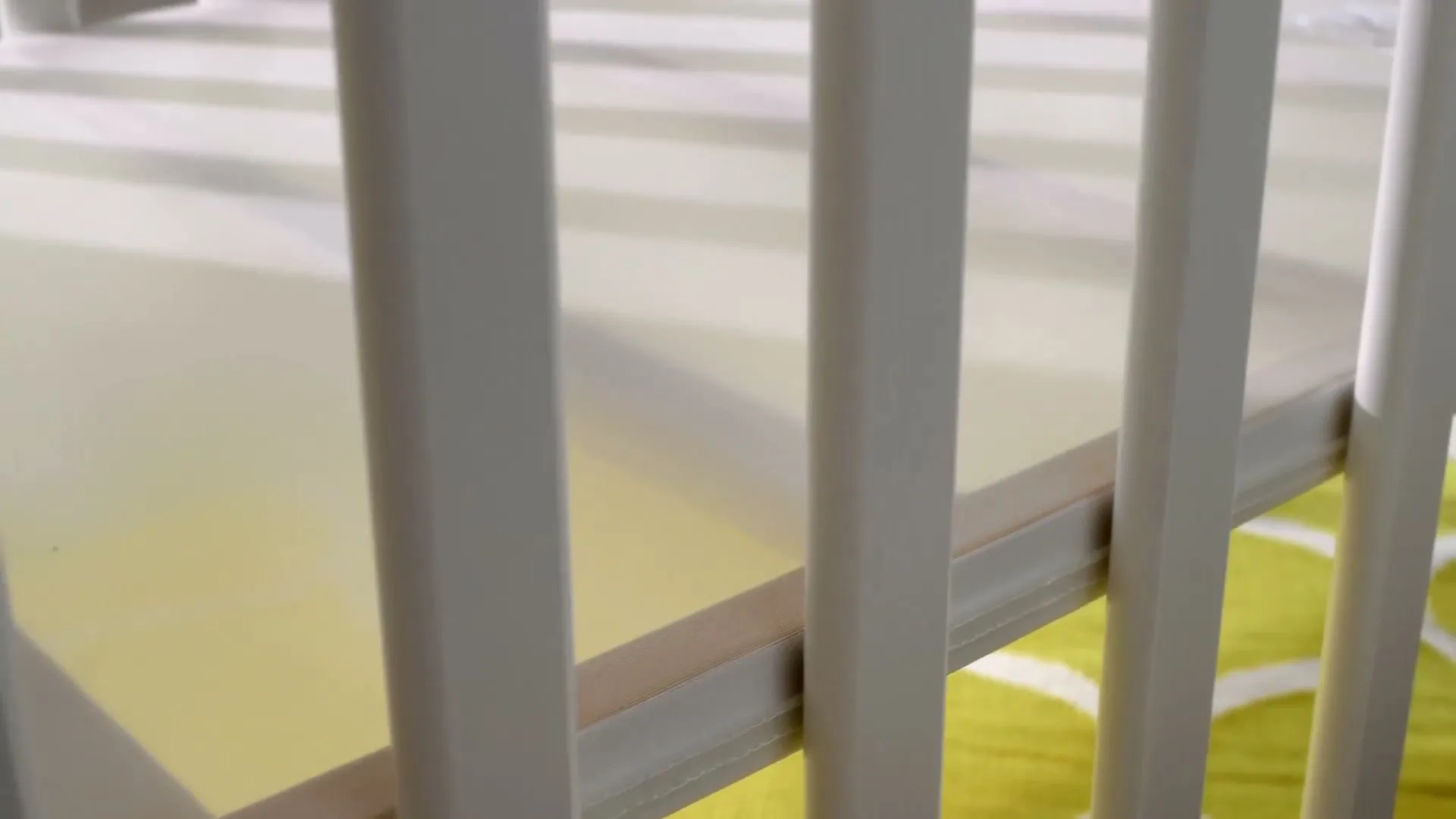 A close-up shot of a white wooden baby crib with a yellow and white patterned sheet, in an interior setting.