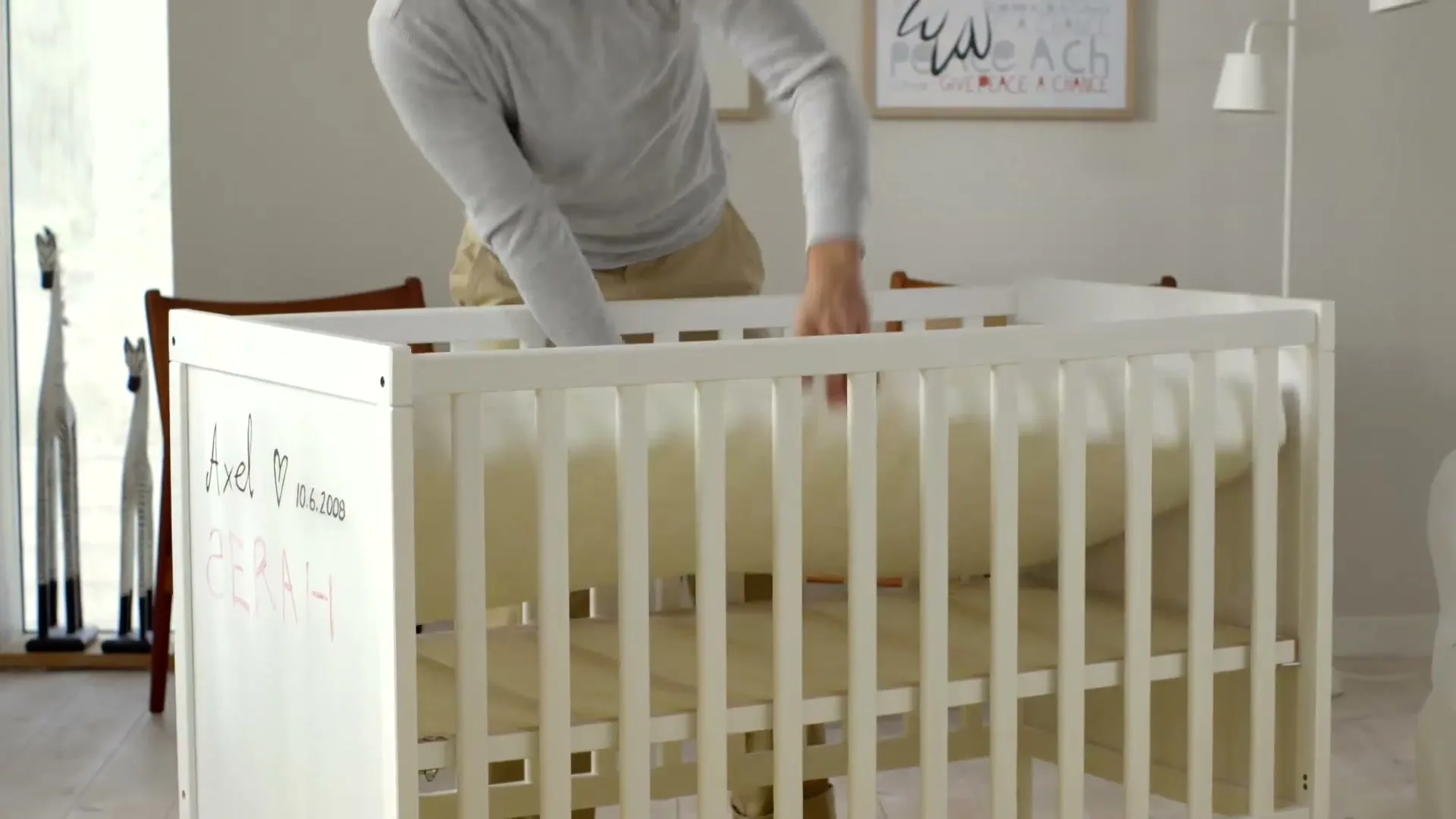 A man in a grey shirt and khaki pants places a white mattress into a white wooden baby crib in a bright, modern room.