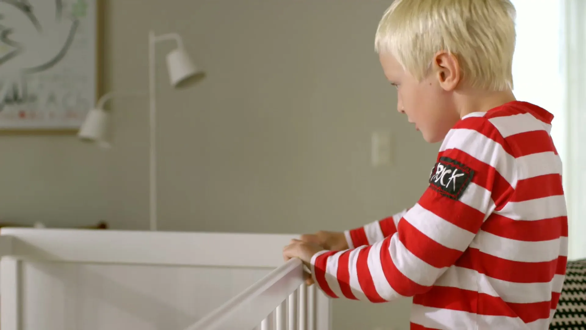 A young boy with blonde hair and a red and white striped shirt looks into a white crib in a bright room.
