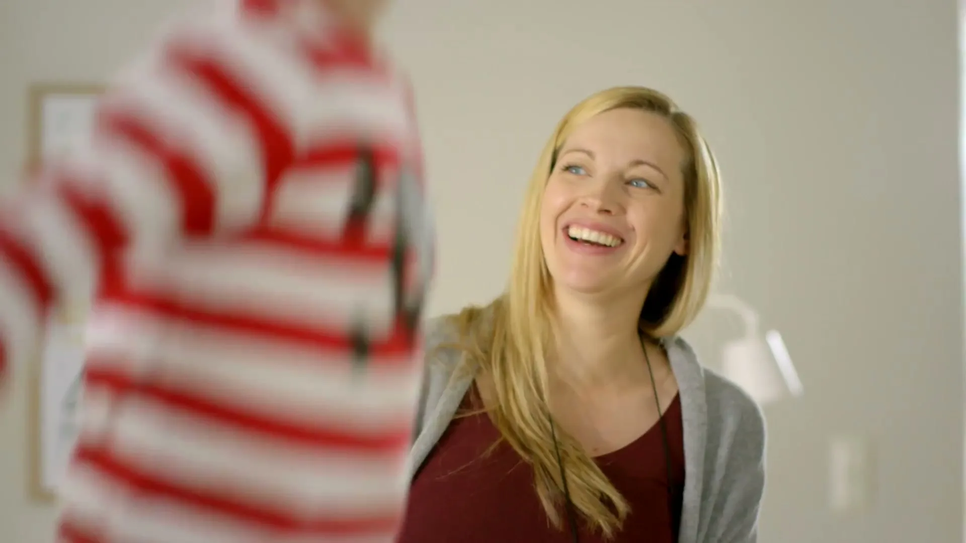 A blonde woman in a maroon top and grey cardigan smiles happily at a child in a red and white striped shirt, who is out of focus in the foreground.