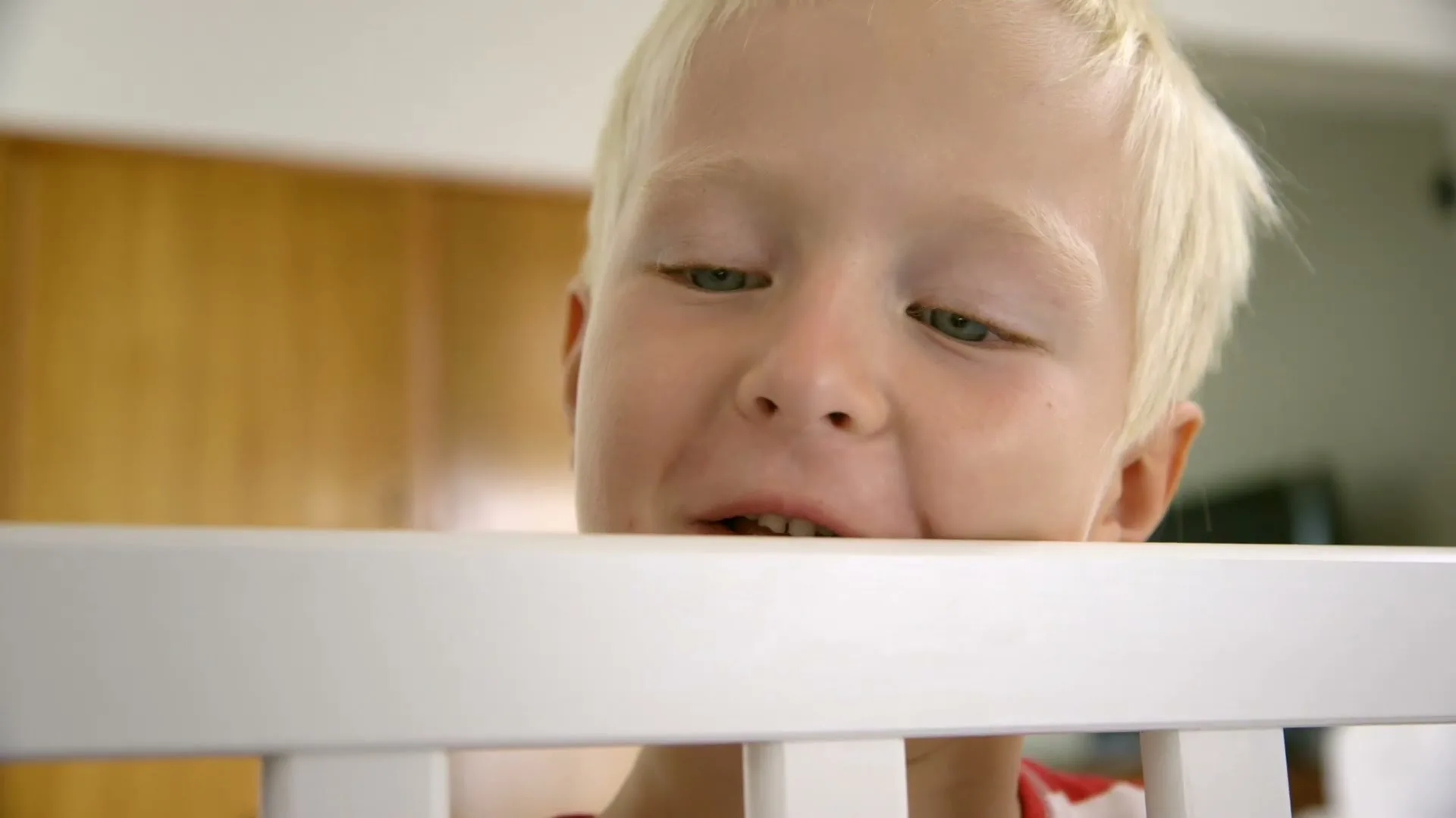 A young blonde boy with blue eyes looks down over the white railing of a crib, smiling slightly in an interior setting.