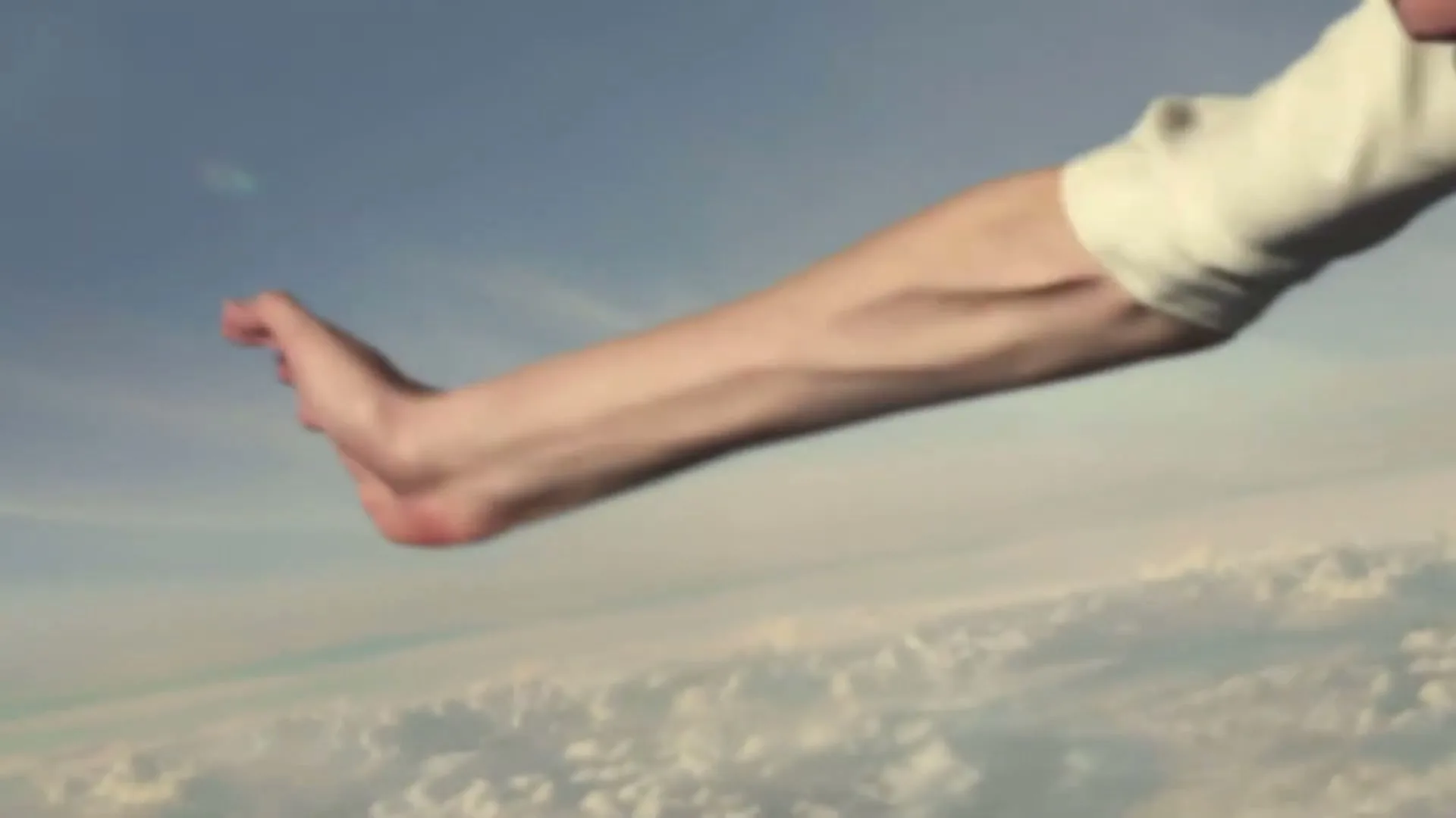 A person's bare arm and foot extend out from a white sheet, seemingly floating above a vast expanse of clouds and blue sky.