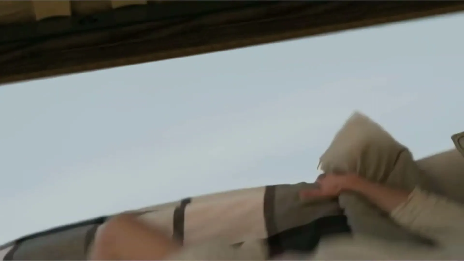 A person's arm and hand are seen pulling a patterned pillow and blanket, with a light blue sky visible in the background.