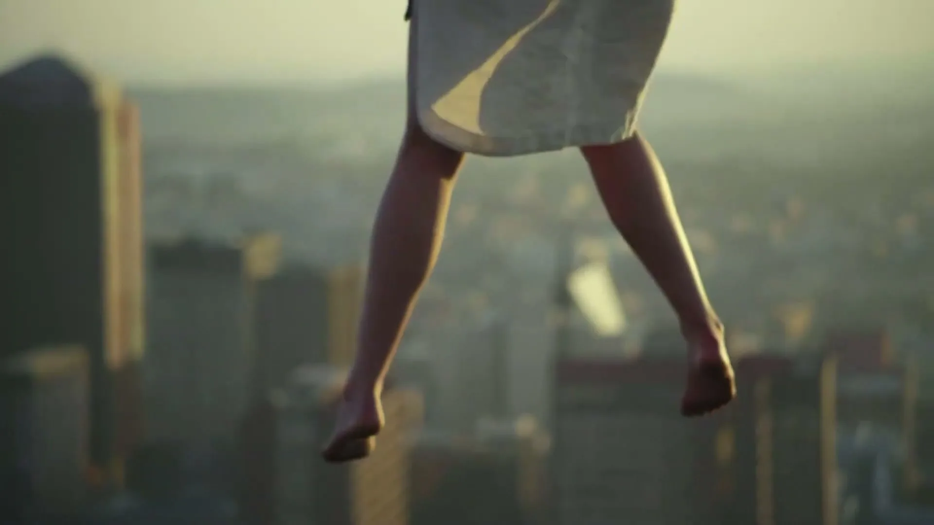 A woman's bare legs dangle in the air, with a city skyline visible far below, suggesting she is floating or falling.