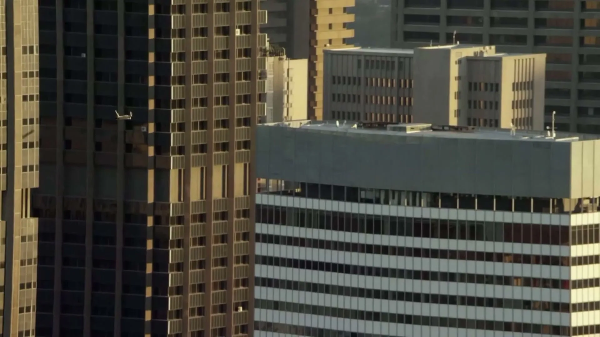 A small white plane flies past tall, modern buildings in a bustling city during the day.