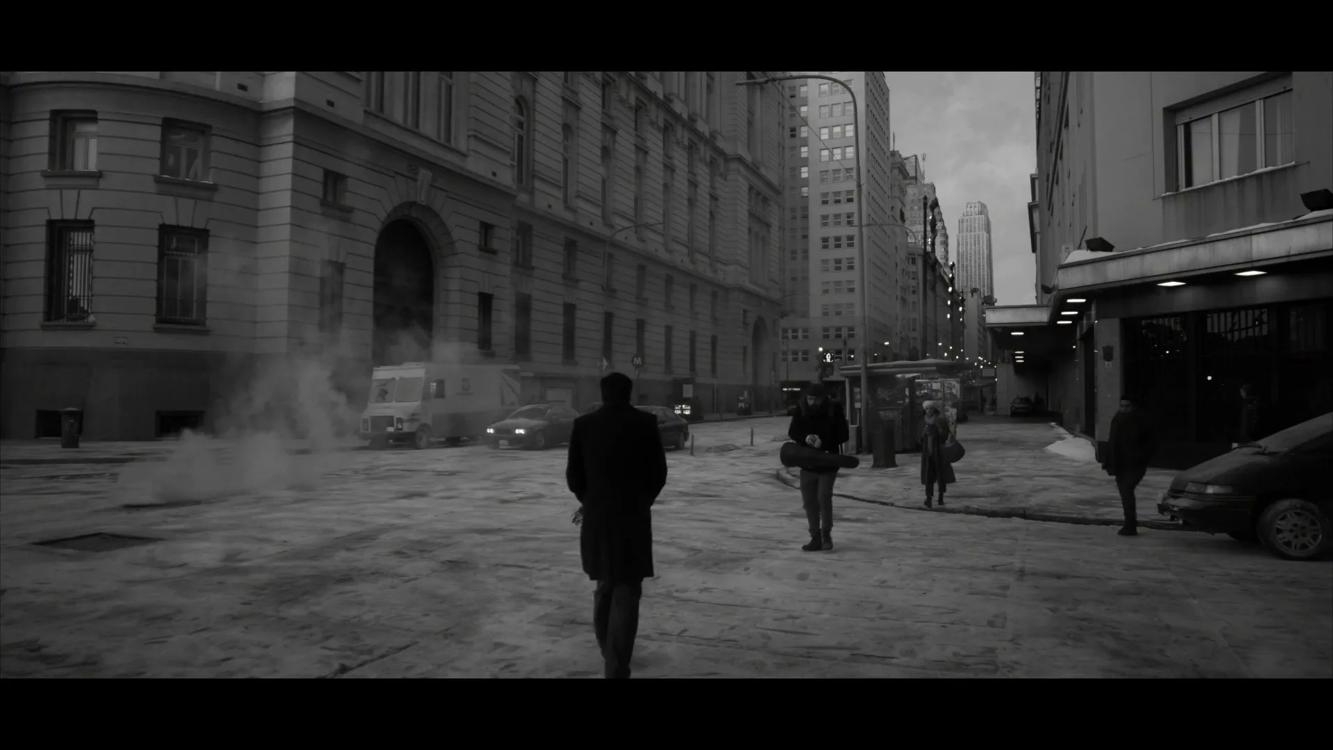 A man in a long coat walks away from the camera down a city street, as other pedestrians walk towards him, in black and white.