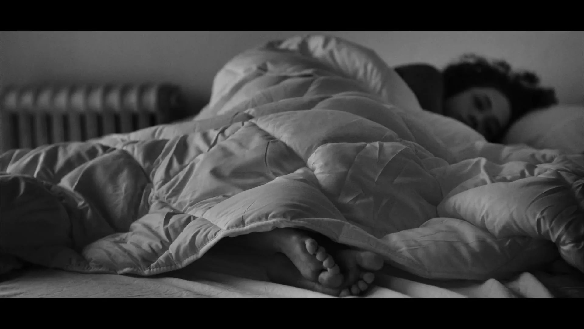 A person with curly hair sleeps soundly under a duvet, with their bare feet peeking out, in a dimly lit bedroom.