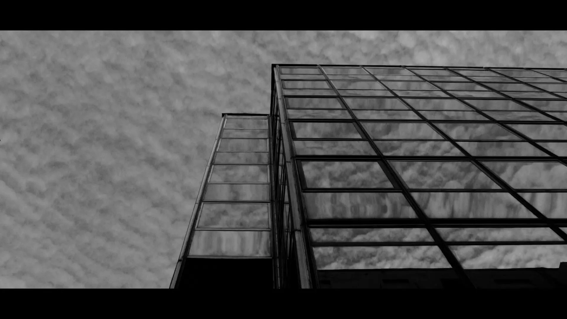 A low-angle, black and white shot of a modern glass building reflecting a cloudy sky, with a subtle camera movement.