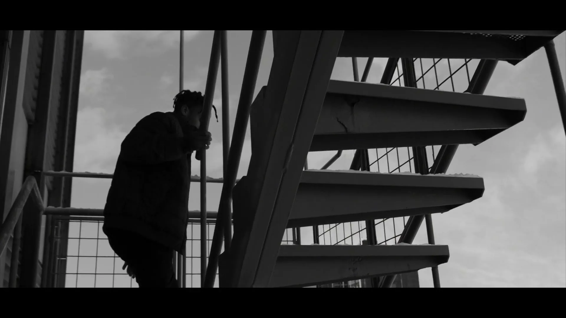 A person with dreadlocks, dressed in a dark jacket, climbs a metal spiral staircase outdoors under a cloudy sky.