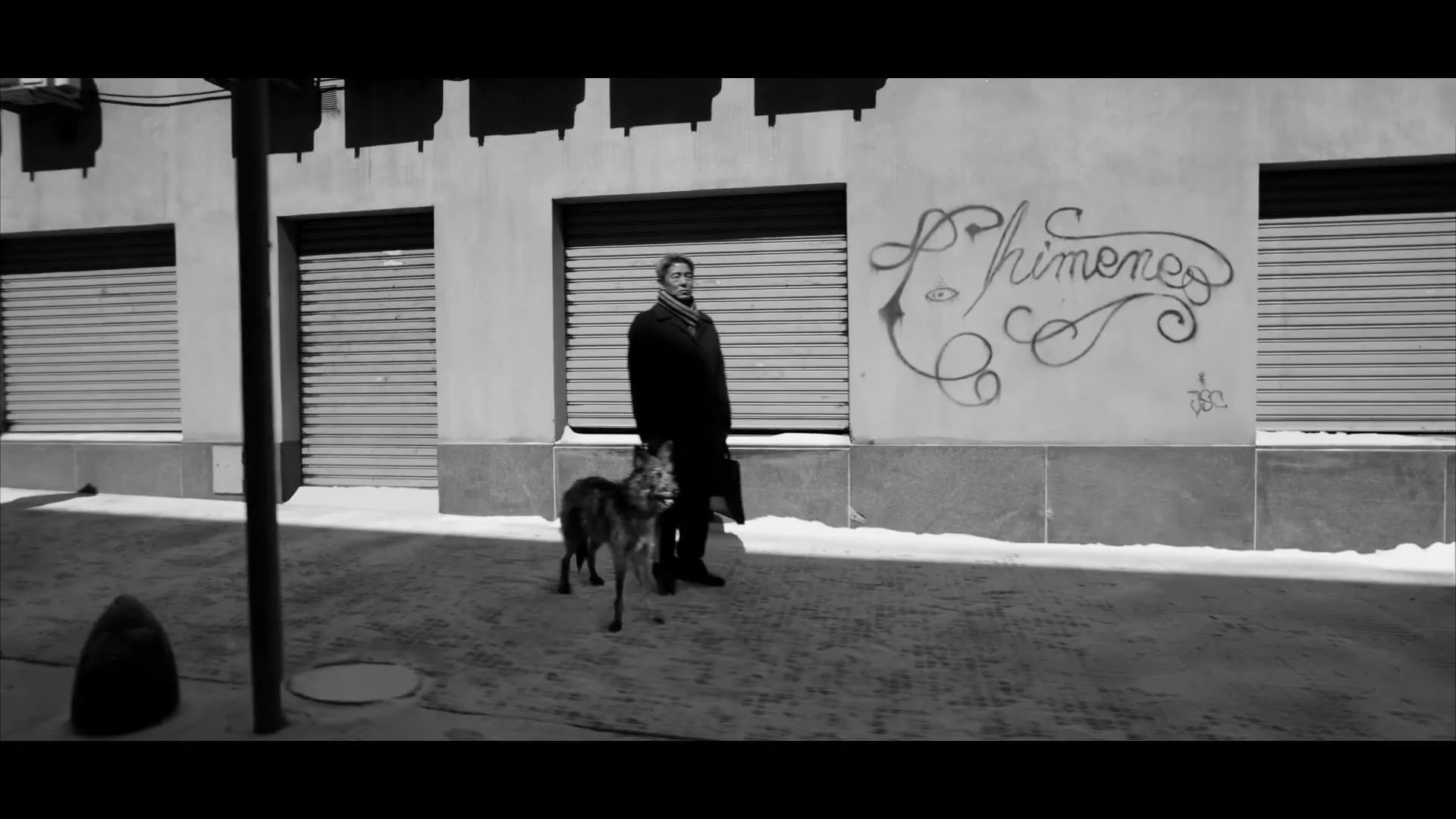 A man in a coat stands with his dog on a snowy street next to a building with graffiti and closed shutters, in black and white.