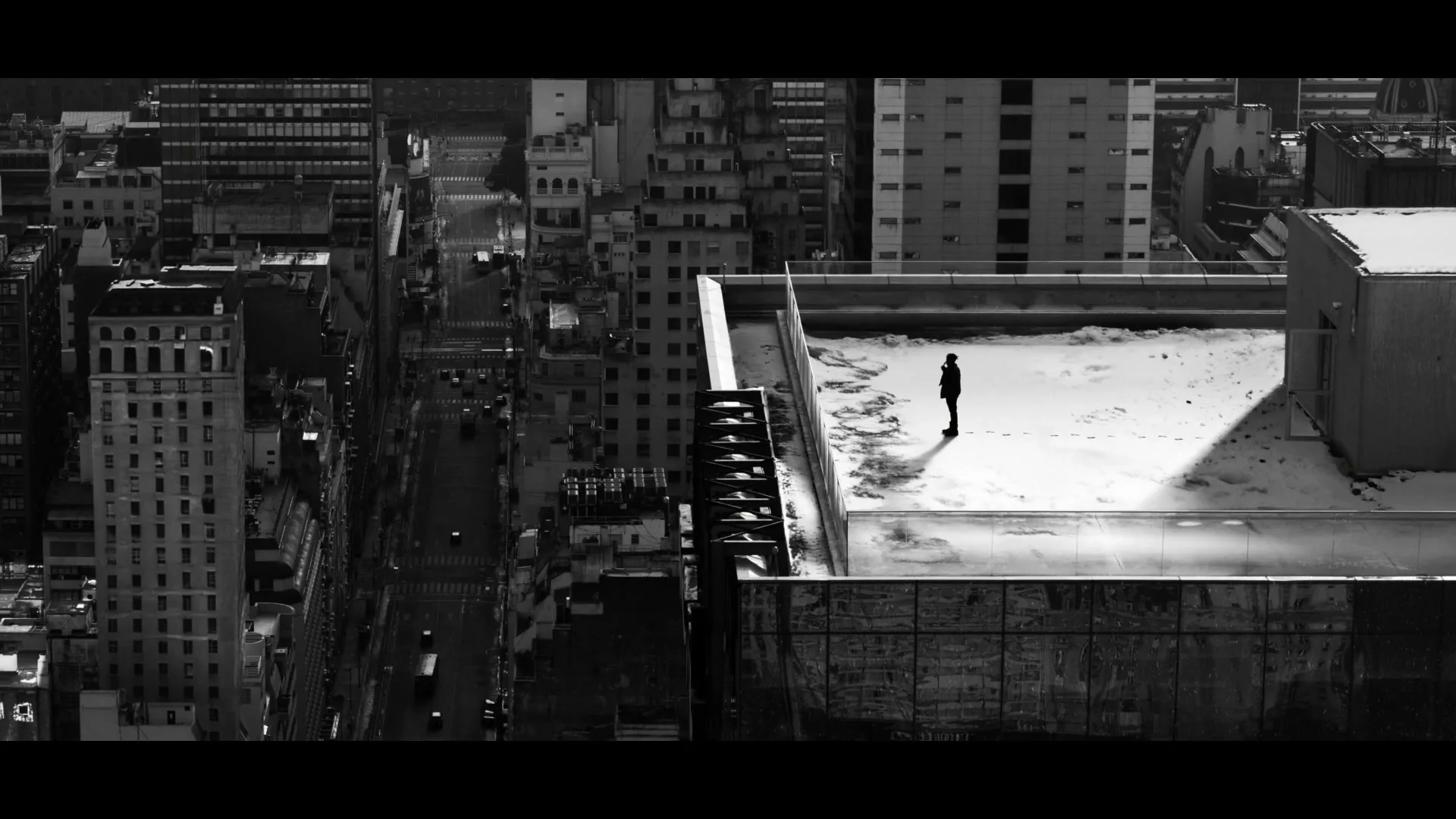 A lone figure stands on a snowy rooftop, looking out over a sprawling city in black and white.