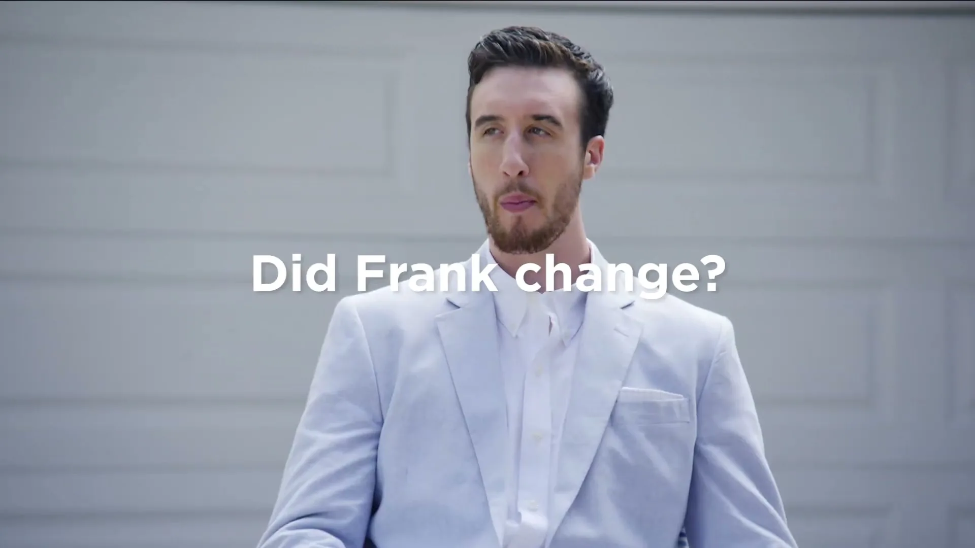 A man in a light blue suit and white shirt sits in front of a garage door, blowing a bubble with gum and looking around.