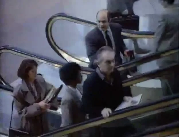 A diverse group of people, including a bald man in a suit, ride escalators up in a busy airport terminal, looking around and talking.
