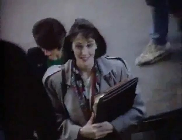 A woman in a trench coat and patterned scarf smiles while holding a folder, surrounded by other people in an airport terminal.