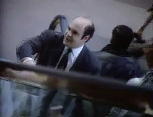 A balding man in a suit and tie, holding onto an escalator handrail, looks up and speaks to someone off-screen in an airport terminal.