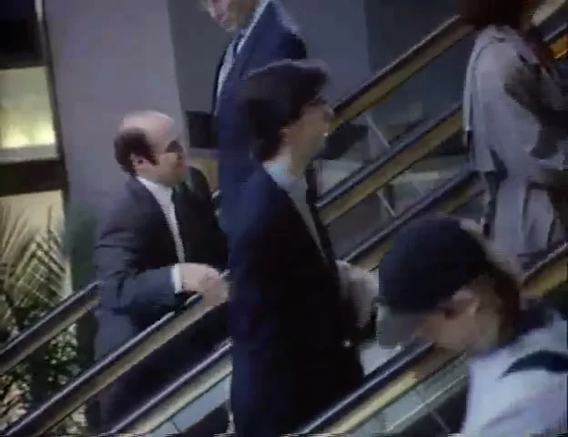 A bald man in a suit on an escalator taps another man on the shoulder and speaks to him, surrounded by other commuters in a modern building.